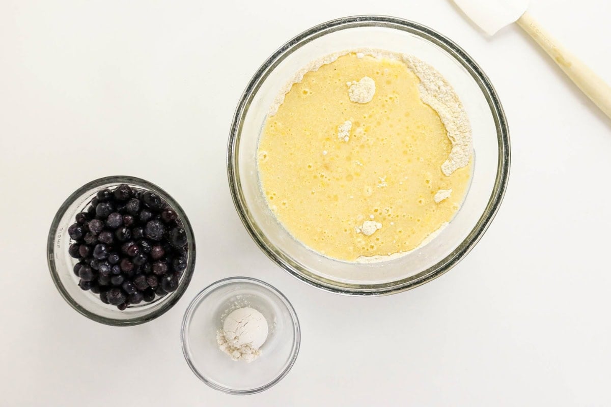 Batter, flour, spatula, and blueberries on a white background.