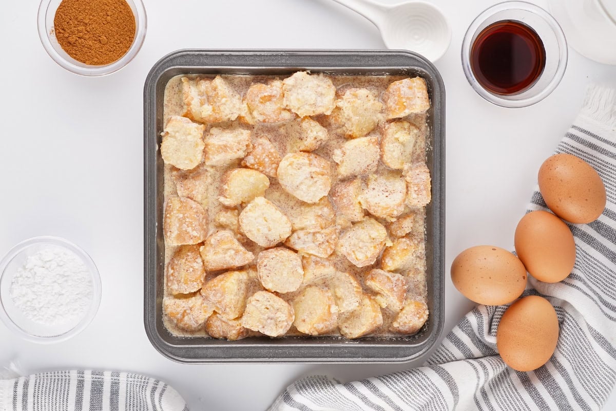 Unbaked bread pudding in a metal pan with eggs and ingredients nearby.