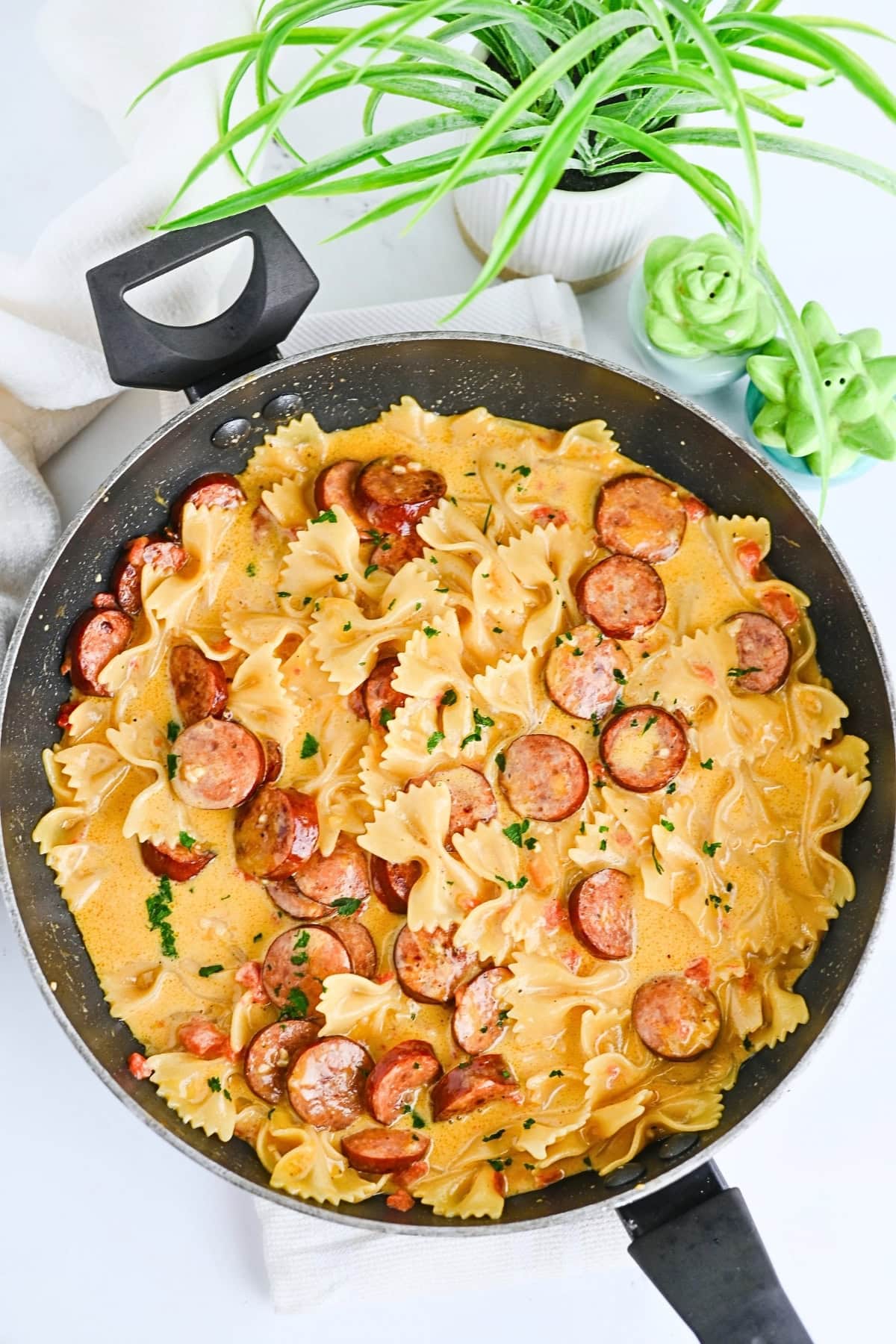 Bowtie pasta with sliced sausage in creamy sauce, in a skillet near plants.