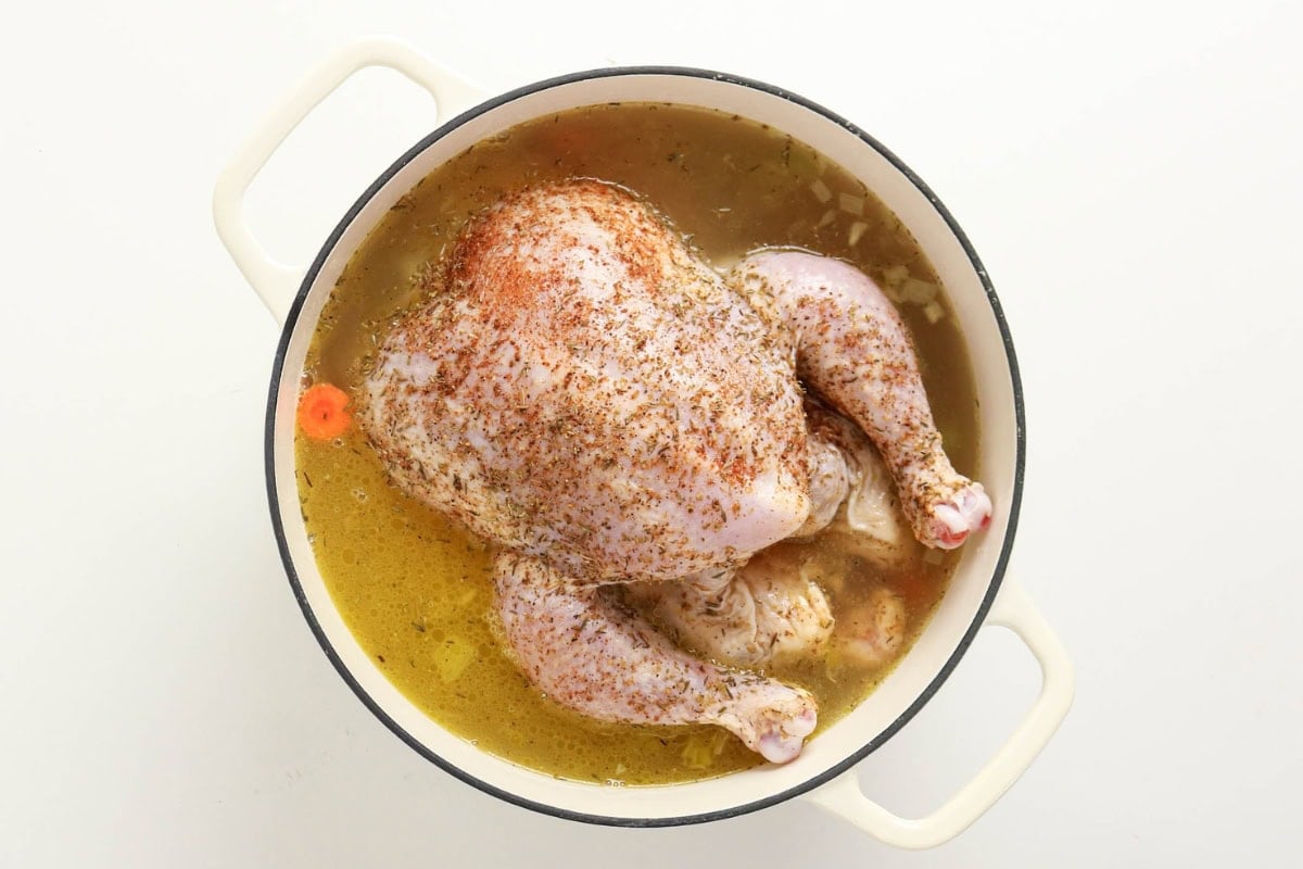 Whole raw chicken in seasoned broth inside a white pot, viewed from above.