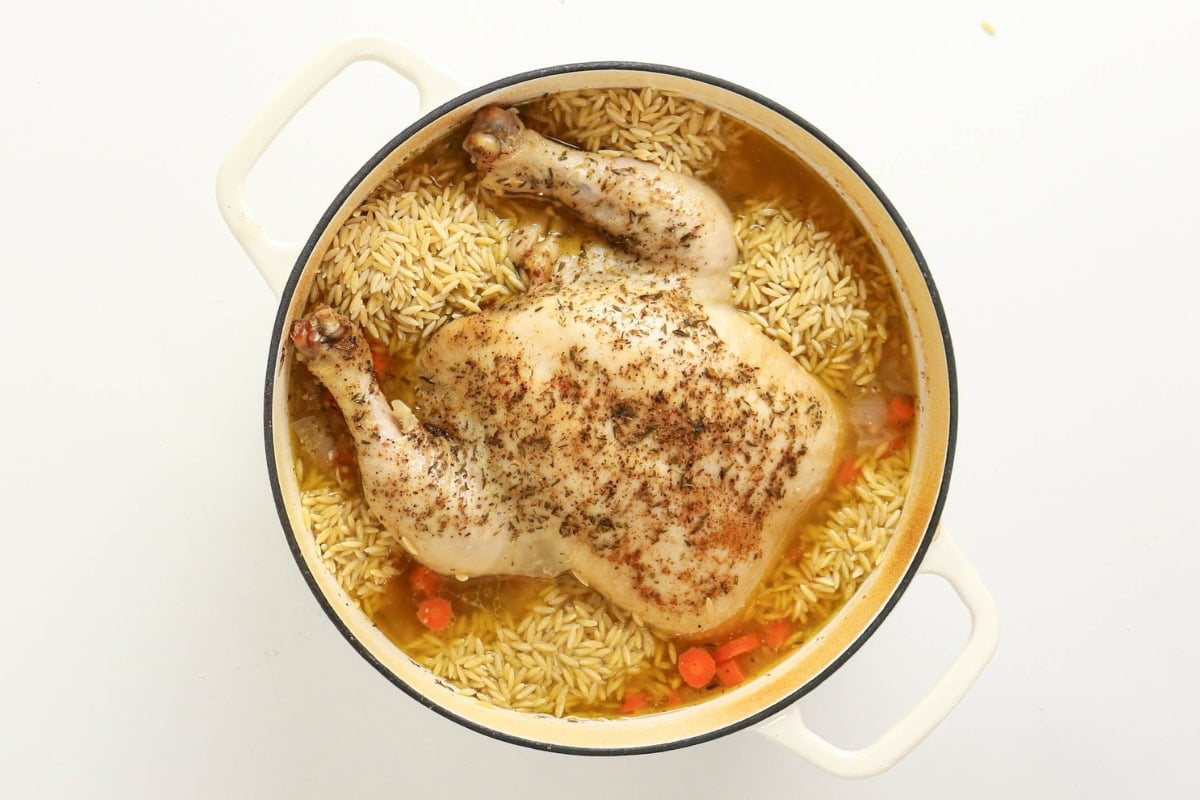Whole seasoned chicken on rice, carrots, and orzo in a white pot.