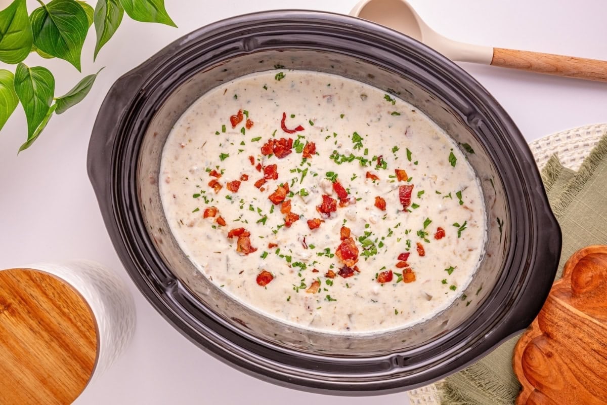 Creamy soup with bacon bits in a slow cooker, garnished with chopped herbs.