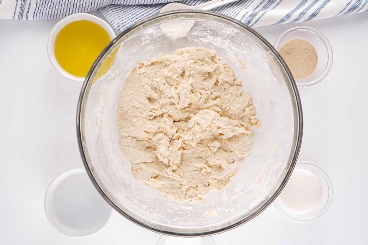 Bowl of bread dough surrounded by small bowls of ingredients on a white surface.