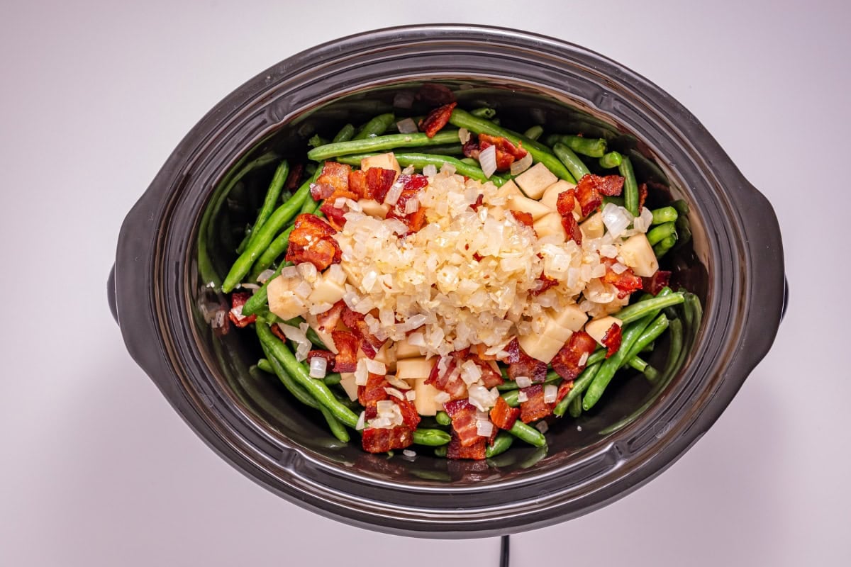 Green beans, cheese, bacon, and onions in a black slow cooker, top view.