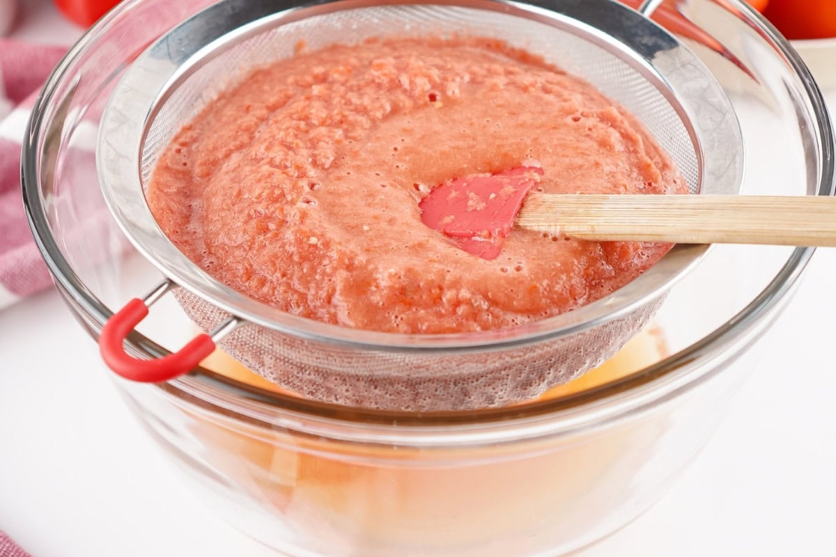 A red spatula strains blended tomatoes through a fine mesh sieve into a bowl.