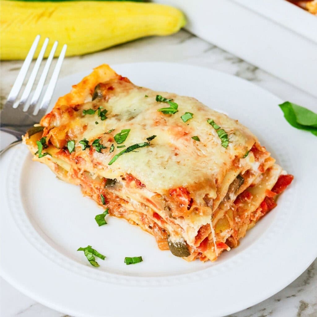 A slice of vegetable lasagna topped with herbs on a white plate with a fork.