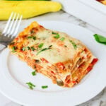 A slice of vegetable lasagna topped with herbs on a white plate with a fork.