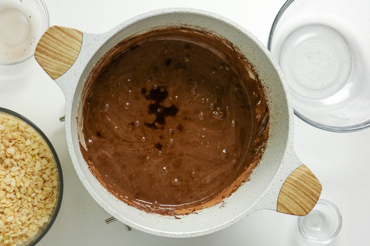 A pot of chocolate mixture sits on a white surface near bowls and ingredients.
