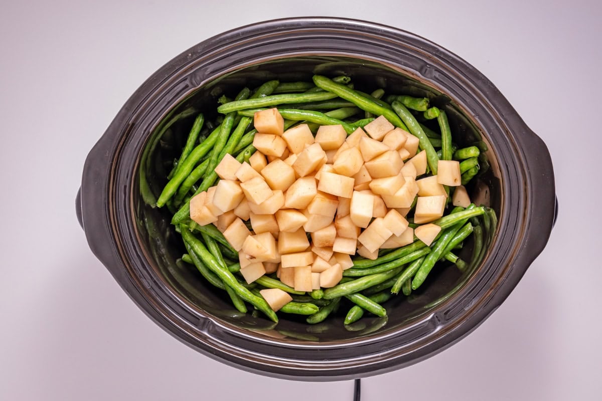 Green beans and cubed potatoes in a black slow cooker, viewed from above.
