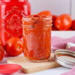 A jar of tomato paste on a wooden board surrounded by fresh tomatoes.
