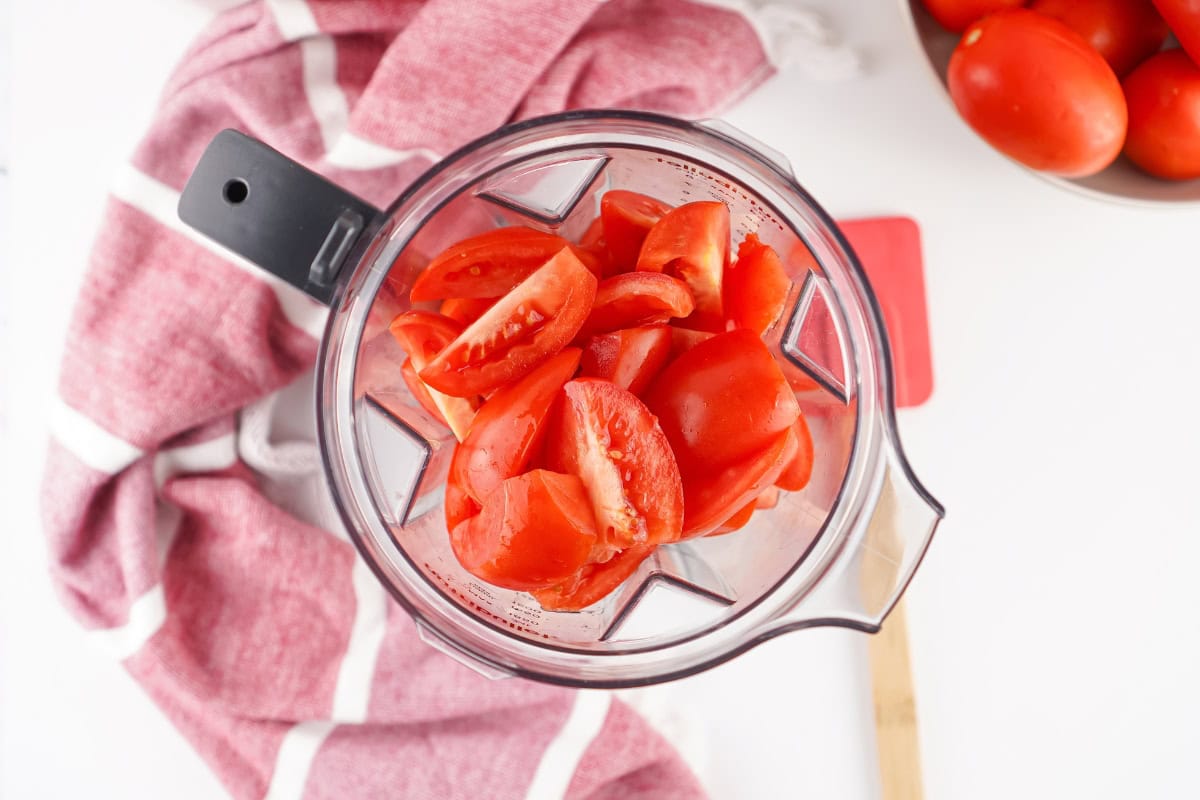 Chopped tomatoes in a blender with a red towel and bowl of tomatoes nearby.