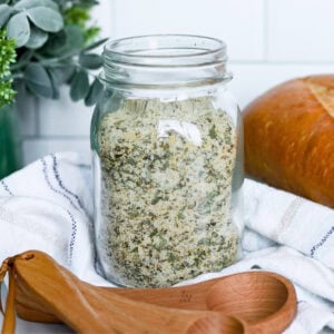 Mason jar of herb seasoning mix beside wooden measuring spoons and bread.