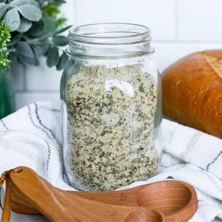 Mason jar of herb seasoning mix beside wooden measuring spoons and bread.