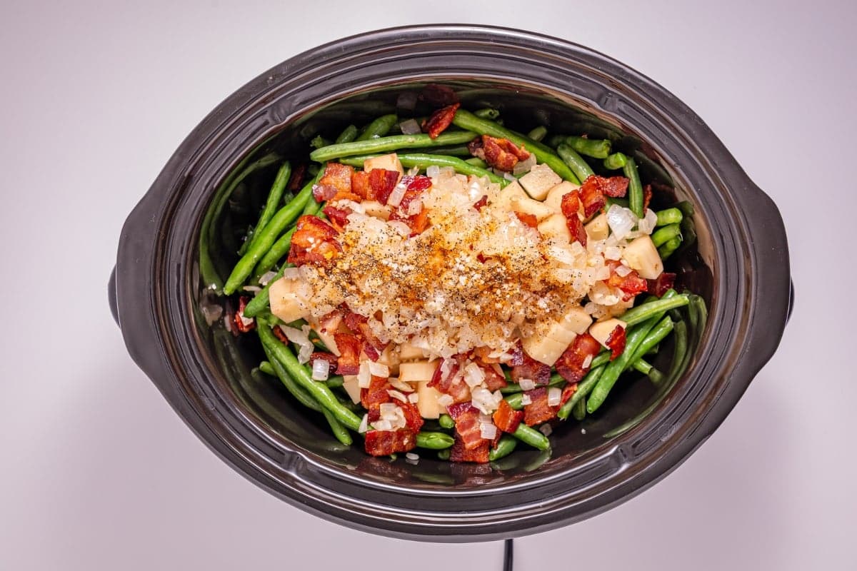 Green beans, bacon, onions, and cheese in a black slow cooker, seen from above.
