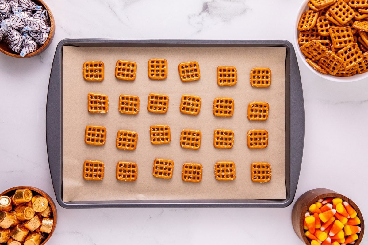 Pretzel squares on a baking sheet surrounded by bowls of snacks and candies.