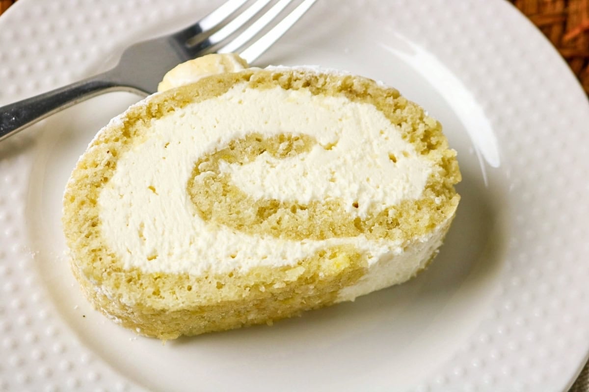 A slice of vanilla cake roll with cream filling on a white plate and fork.