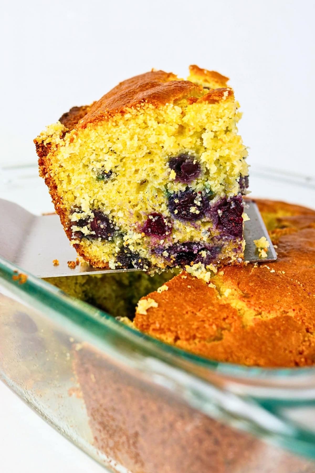 A slice of blueberry cornbread being lifted from a glass baking dish.