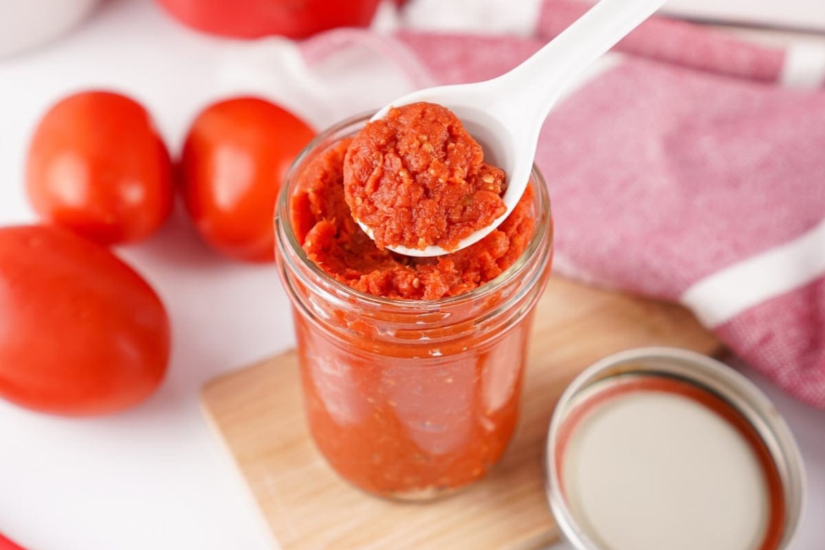 A spoonful of tomato paste held above a glass jar with tomatoes nearby.