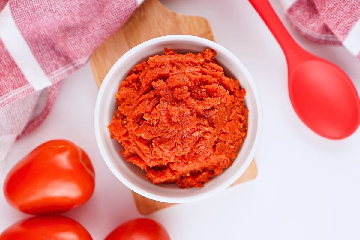 A bowl of tomato paste with fresh tomatoes and a red spoon nearby.
