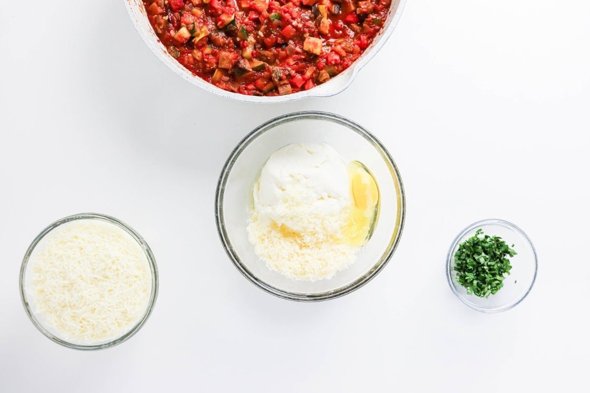 Tomato sauce, ricotta mix, shredded cheese, and chopped herbs in bowls on white.