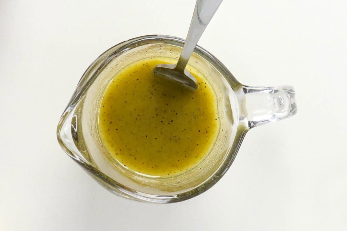 Glass pitcher with yellow salad dressing and a metal spoon on white background.