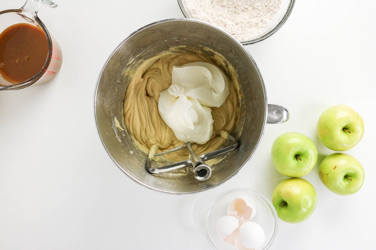 Cake batter with sour cream, apples, eggs, flour, and caramel on a counter.