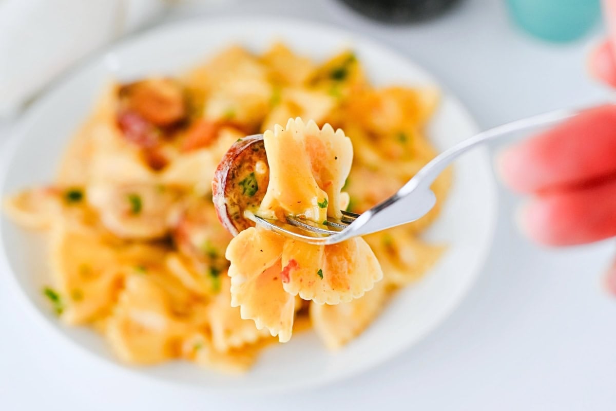 Fork holding bowtie pasta with sauce and herbs, plate of pasta in background.