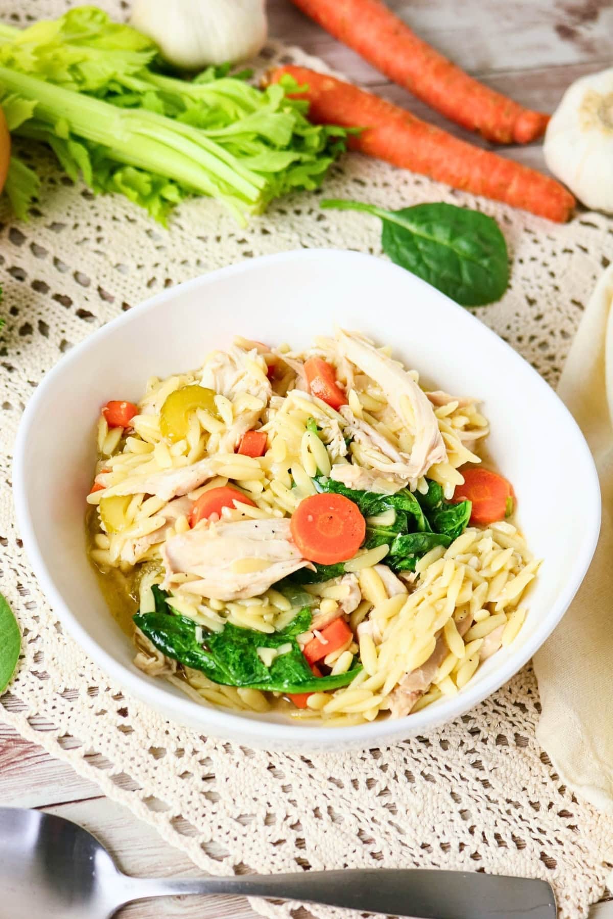 Bowl of chicken soup with orzo, carrots, celery, and spinach on a lace mat.