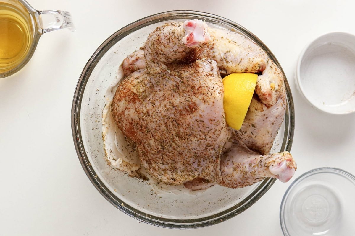 Raw seasoned whole chicken with lemon wedge in a glass bowl on a white surface.