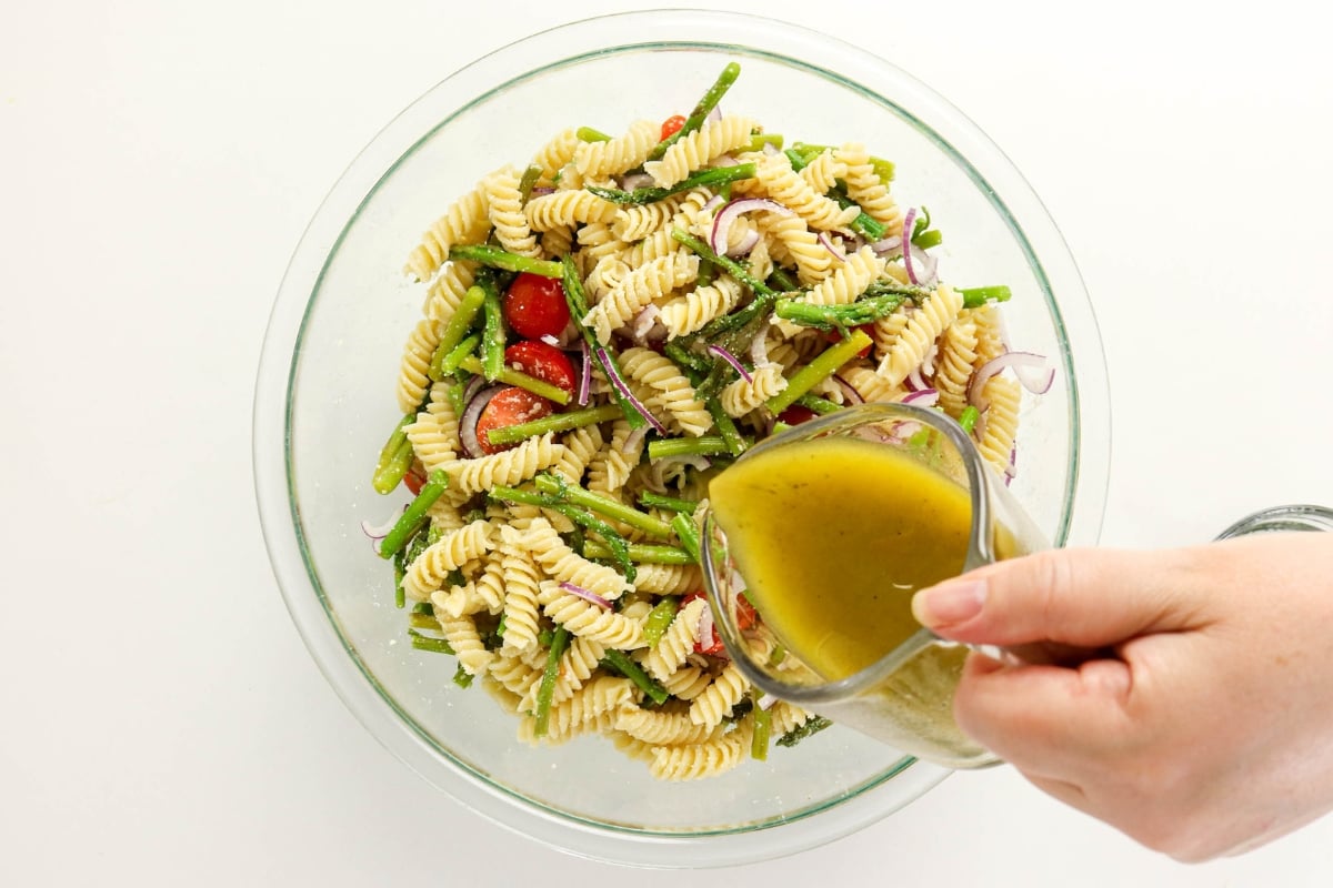Hand pouring dressing onto pasta salad with tomatoes, asparagus, and red onion.