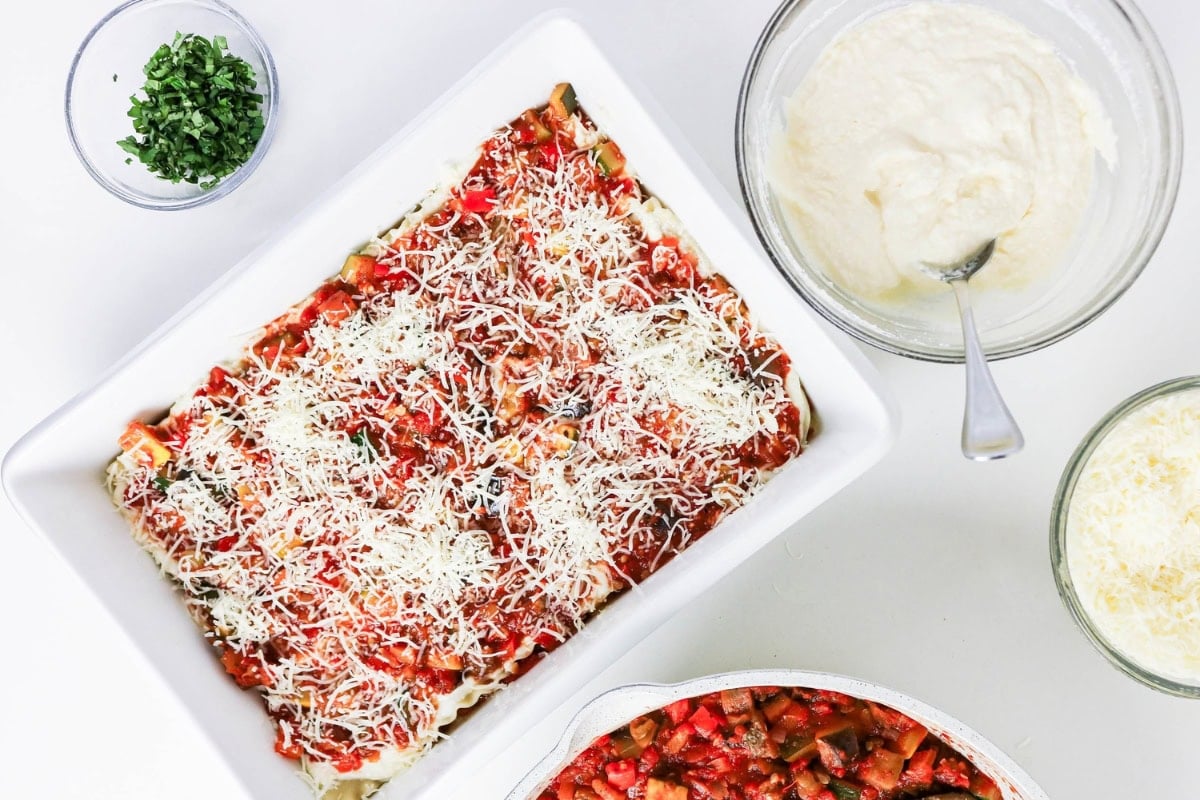 Unbaked lasagna in a dish, with bowls of sauce, cheese, and herbs beside it.
