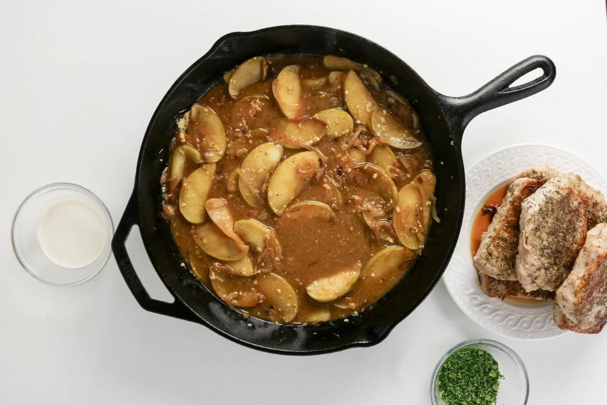 Sliced apples in sauce in a skillet, with pork chops on a plate nearby.