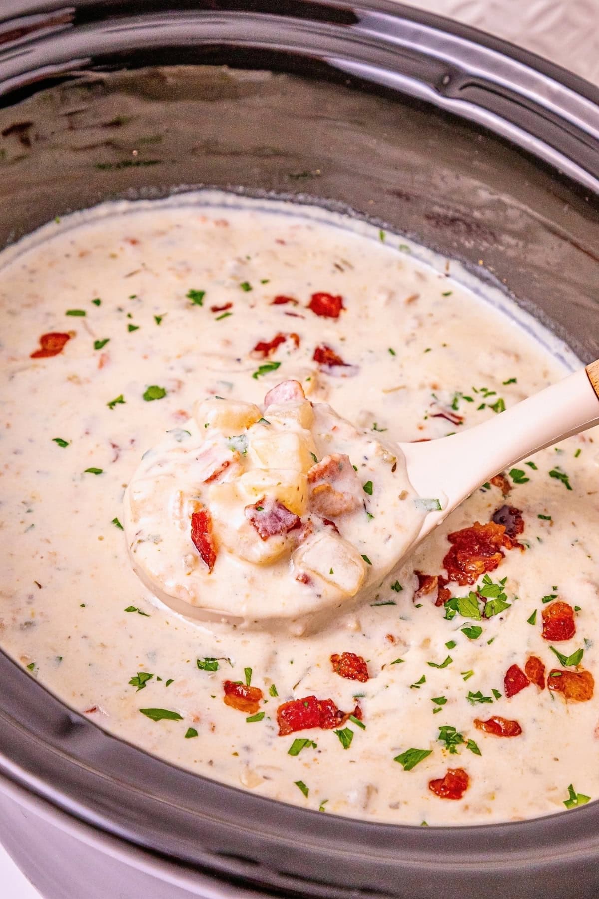Creamy soup with bacon, potatoes, and herbs in a ladle over a crockpot.