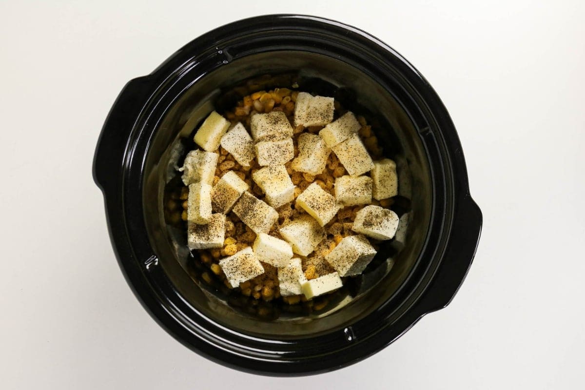 Cubed butter, cheese, and seasoning on pasta in a black slow cooker.