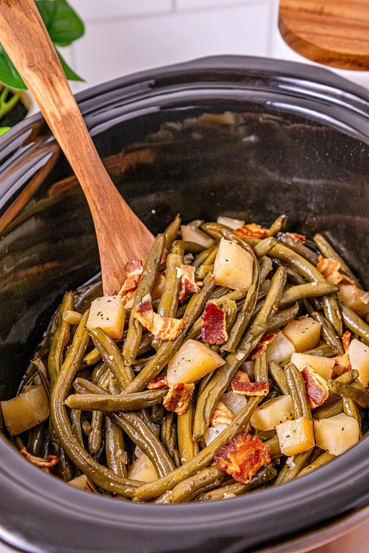 Slow-cooked green beans with potatoes and bacon in a black crockpot.