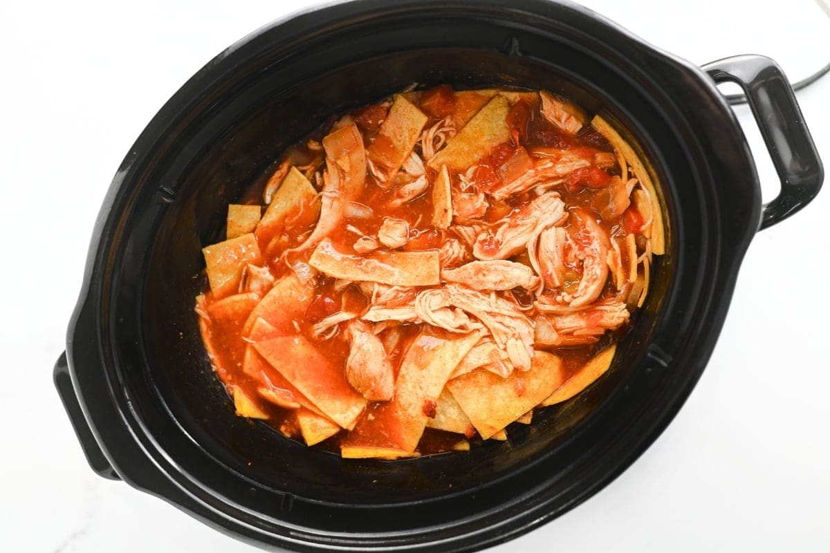 Shredded chicken and tortilla strips in red sauce inside a slow cooker.