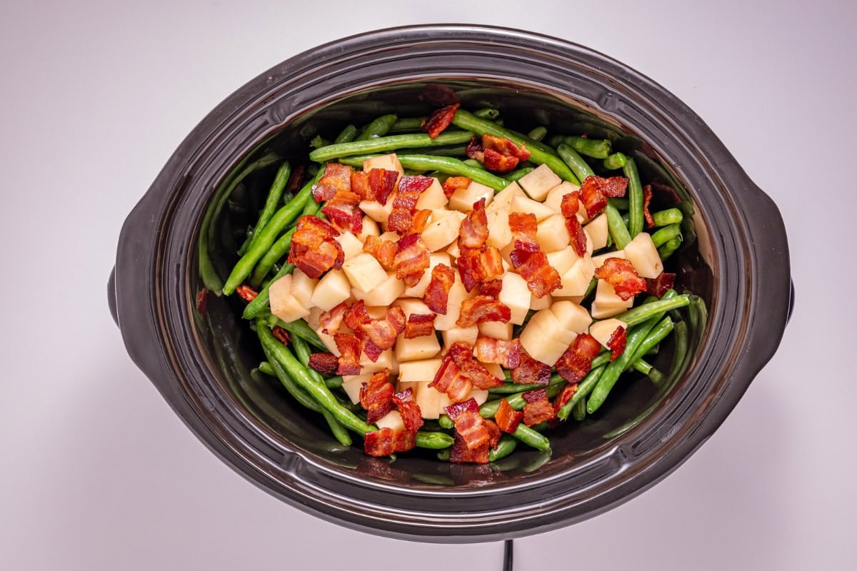 Slow cooker with green beans, diced potatoes, and crispy bacon on top.