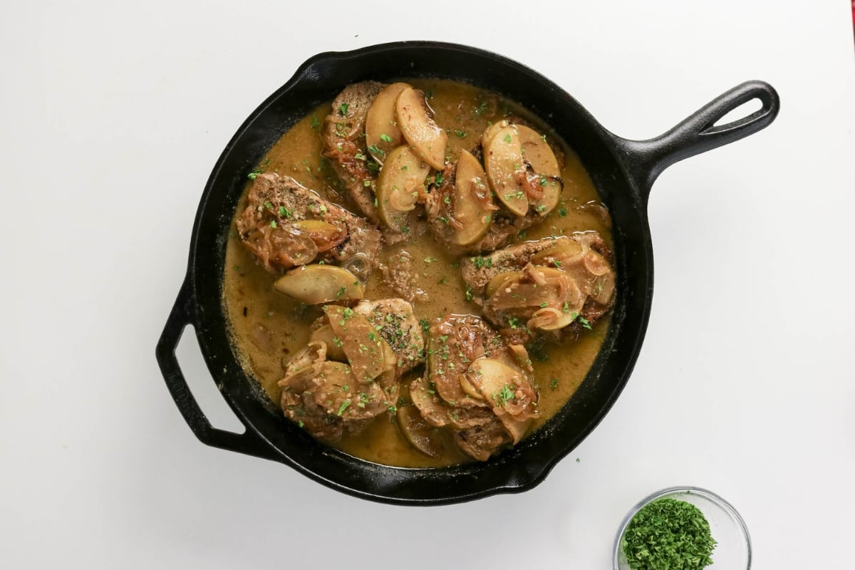 Chicken thighs with apples and sauce in a black skillet, garnished with herbs.