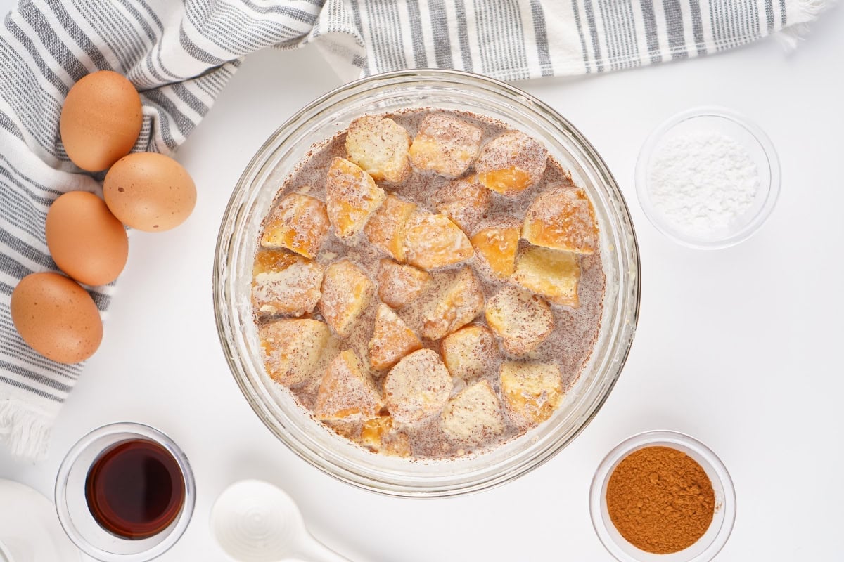 Bowl of bread soaking in custard with eggs, cinnamon, and sugar on the side.