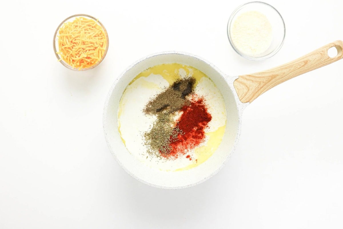 White pan with cream, spices, and cheese; two bowls of cheese beside it.