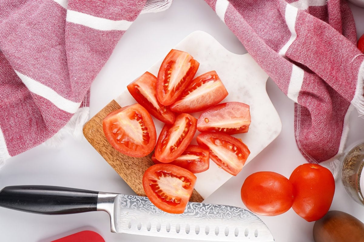 Tomato wedges on a cutting board with a knife and red kitchen towels nearby.