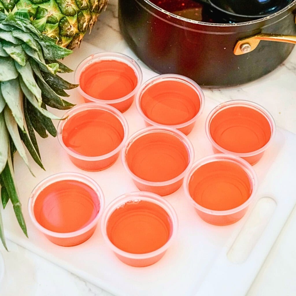 Eight small plastic cups filled with pink gelatin next to a whole pineapple.