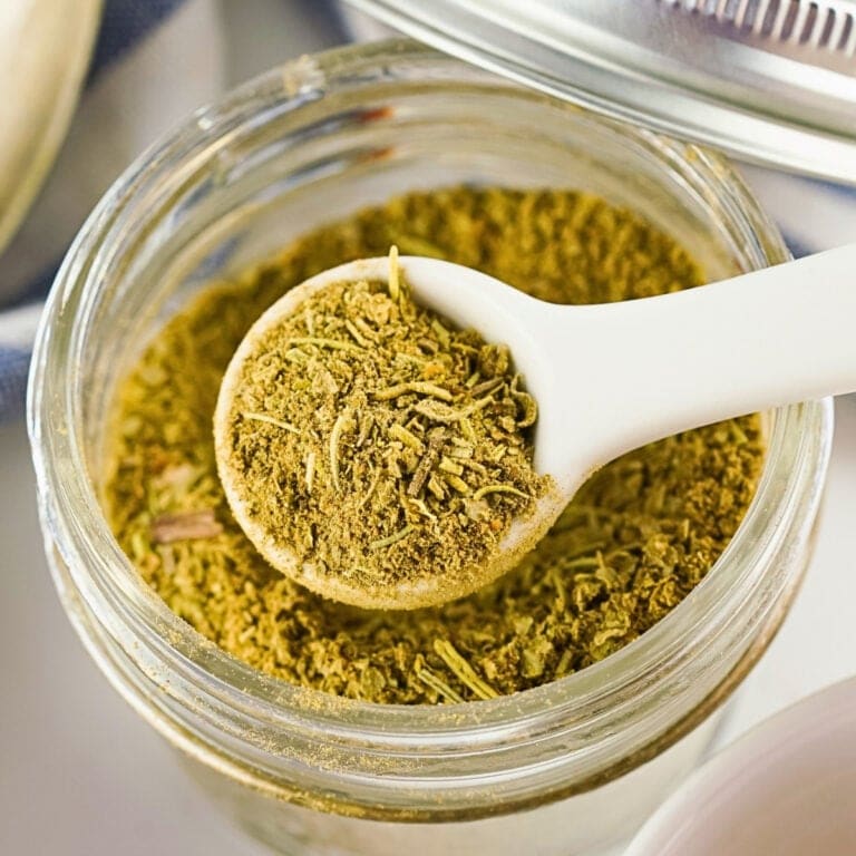 A white spoon holding green dried herbs over a glass jar of the same spice.