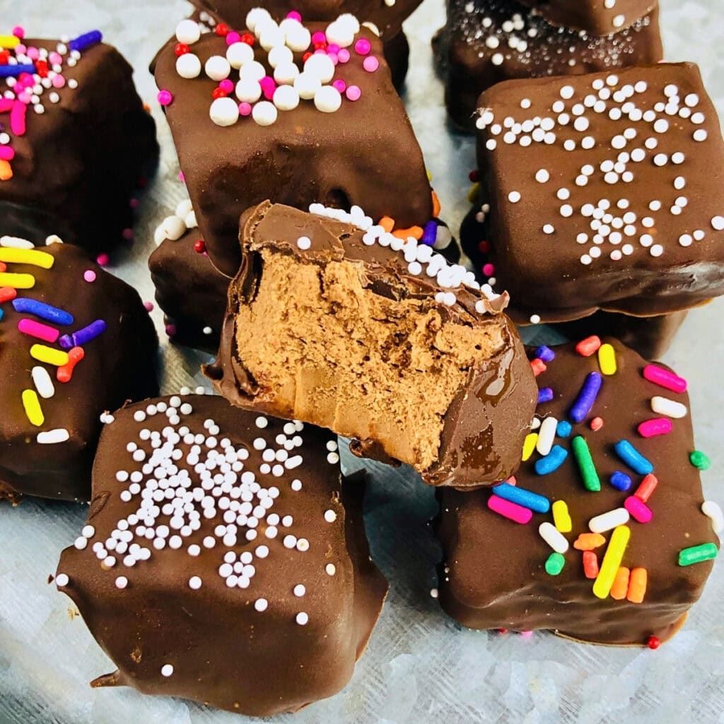 Chocolate-covered squares with colorful sprinkles, one has a bite taken out.