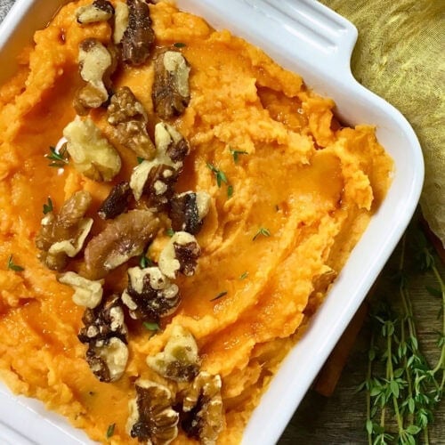 Mashed sweet potatoes topped with walnuts and fresh thyme in a white dish.
