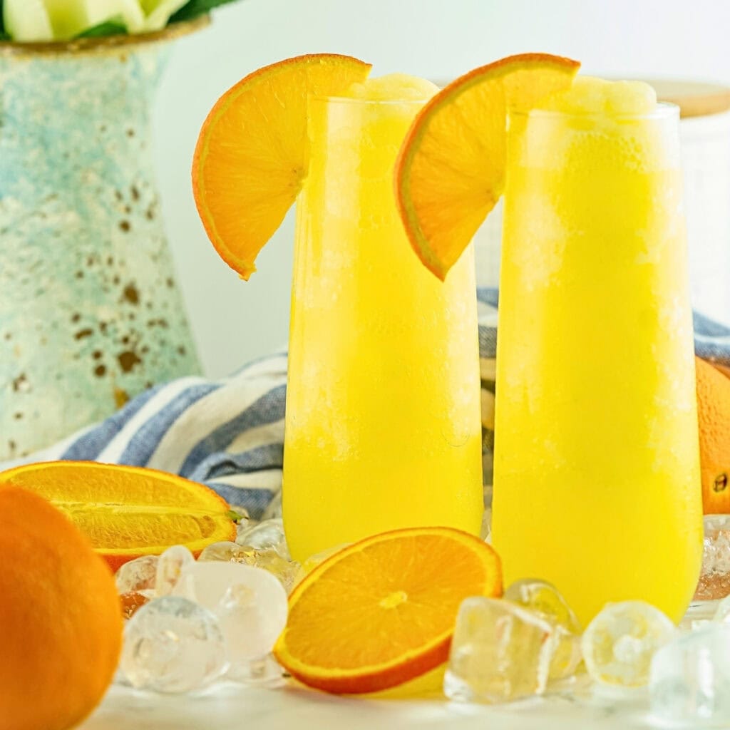 Two tall glasses of orange drink with orange slices and ice cubes nearby.