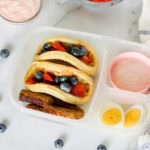 Lunchbox with mini pancakes, berries, boiled egg, yogurt, and banana slices.