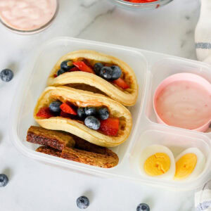 Lunchbox with mini pancakes, berries, boiled egg, yogurt, and banana slices.