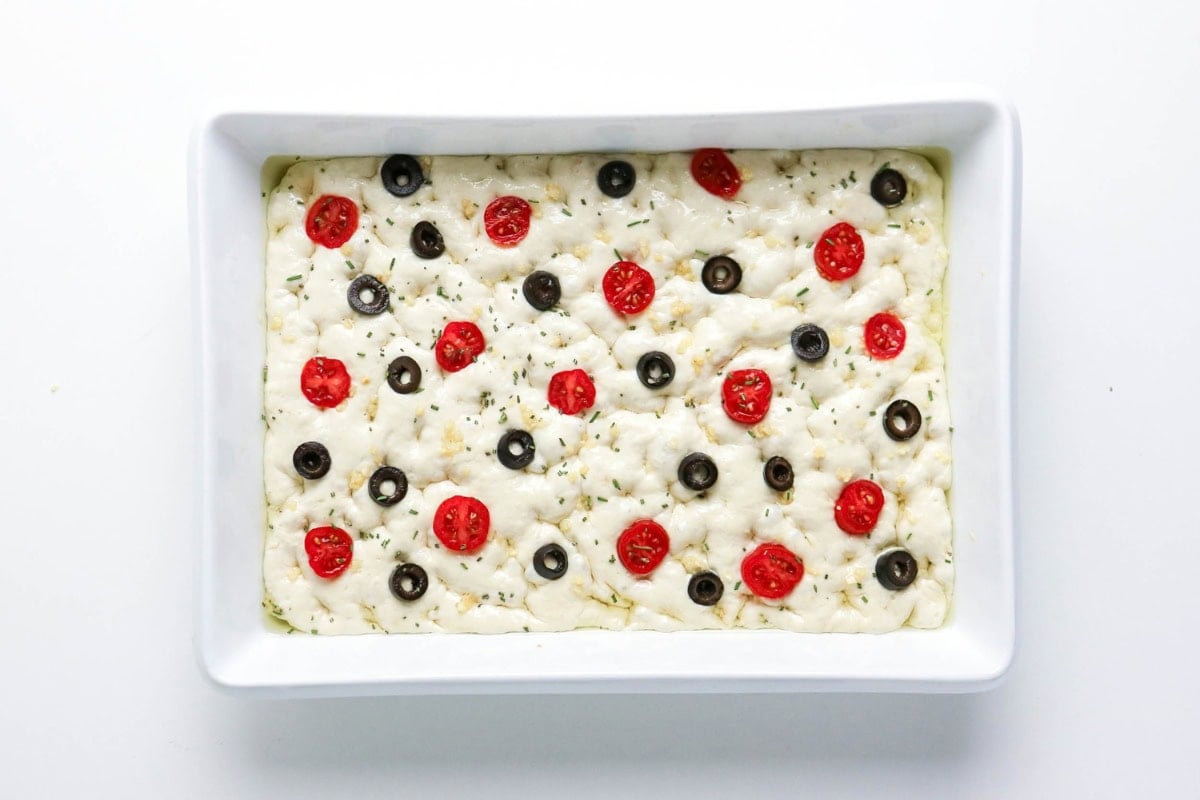 Unbaked focaccia topped with sliced tomatoes and black olives in a white dish.