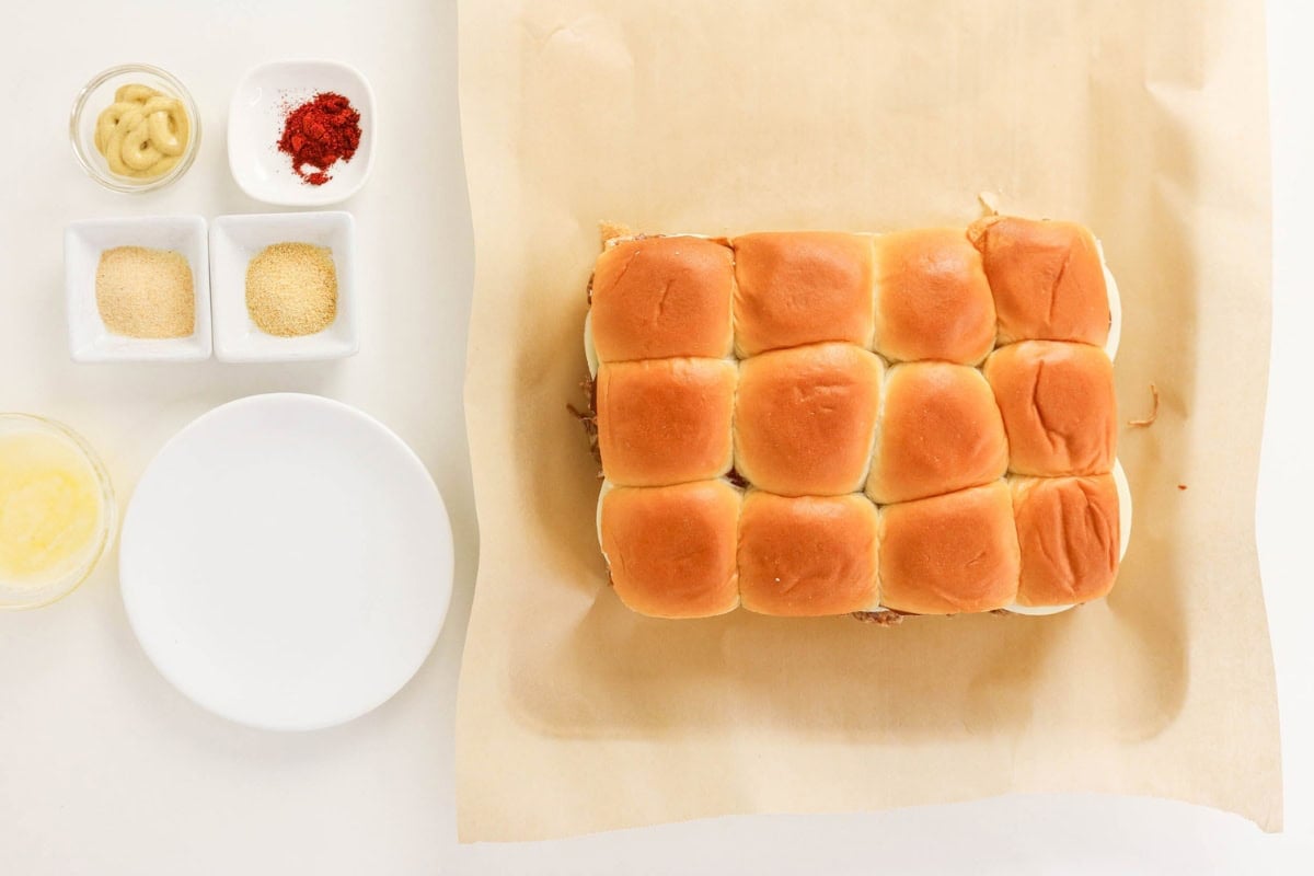 A tray of slider buns on parchment, with seasonings and plates beside it.